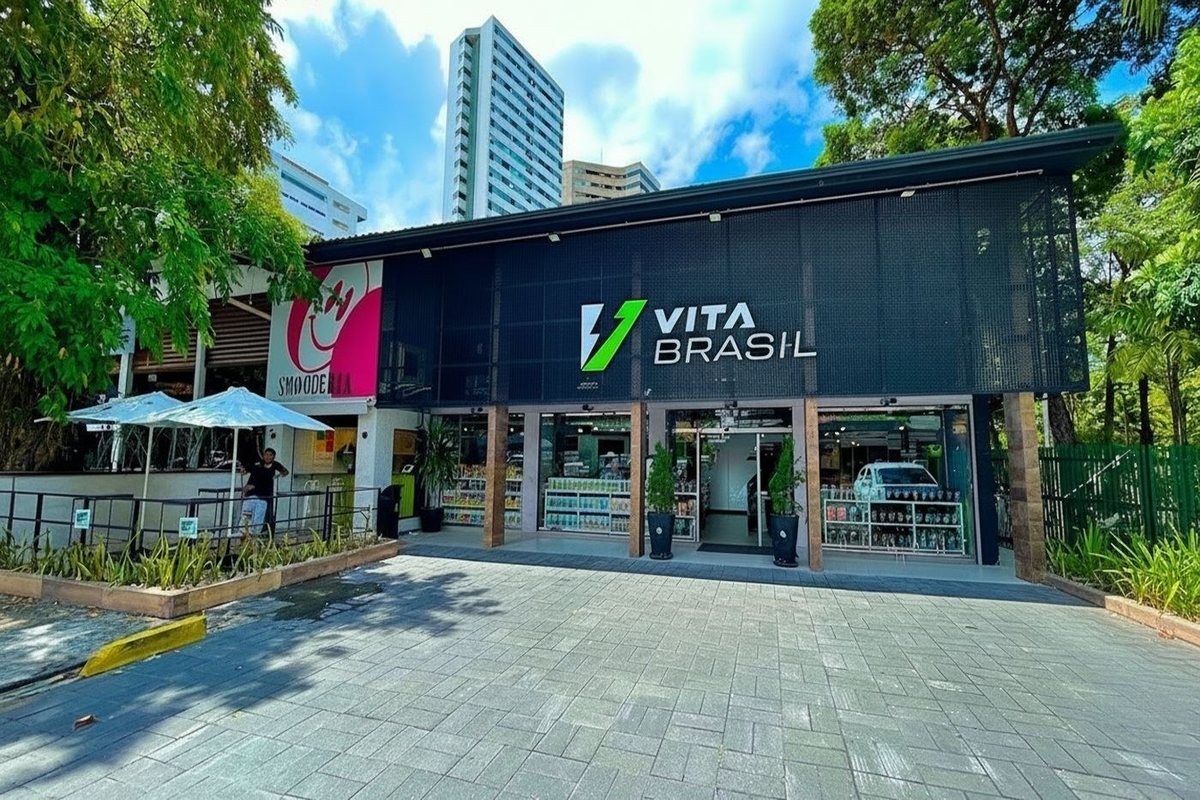 Fachada de uma loja VitaBrasil vista da praça: estrutura escura sob árvores, calçada larga e vitrines abertas com prateleiras ao fundo.